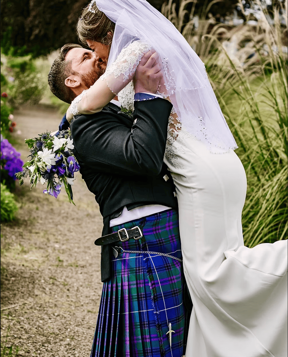 Groom in tartan kilt lifts bride for a kiss along a garden path